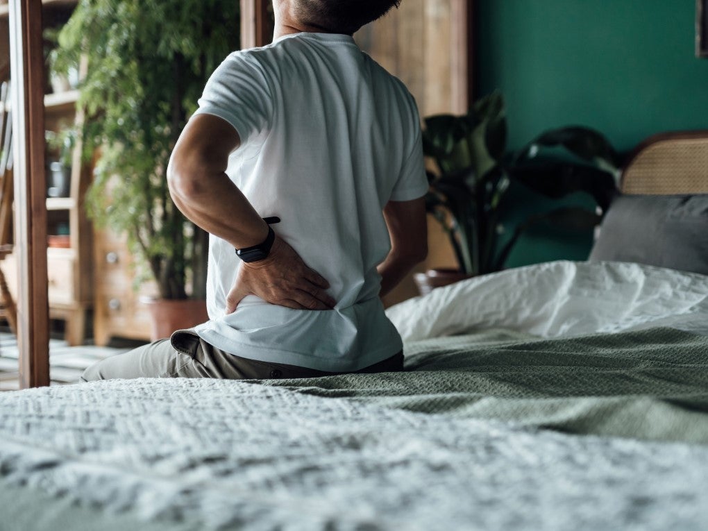 Man facing away from camera sitting on bed while grasping at lower back in pain