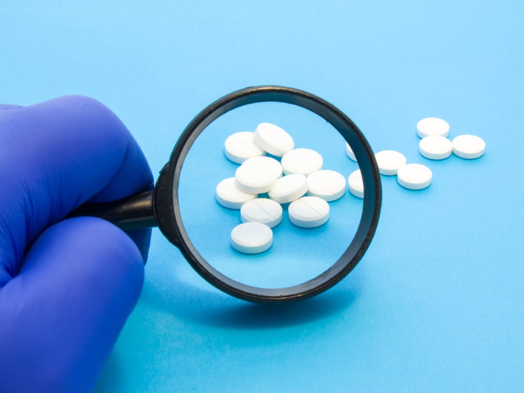 Gloved hand holding a magnifying glass looking at white round pills on a blue background