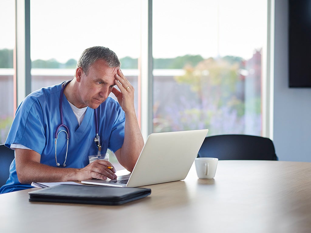 Doctor in scrubs sitting at his desk looking at his computer holding his head in his hands